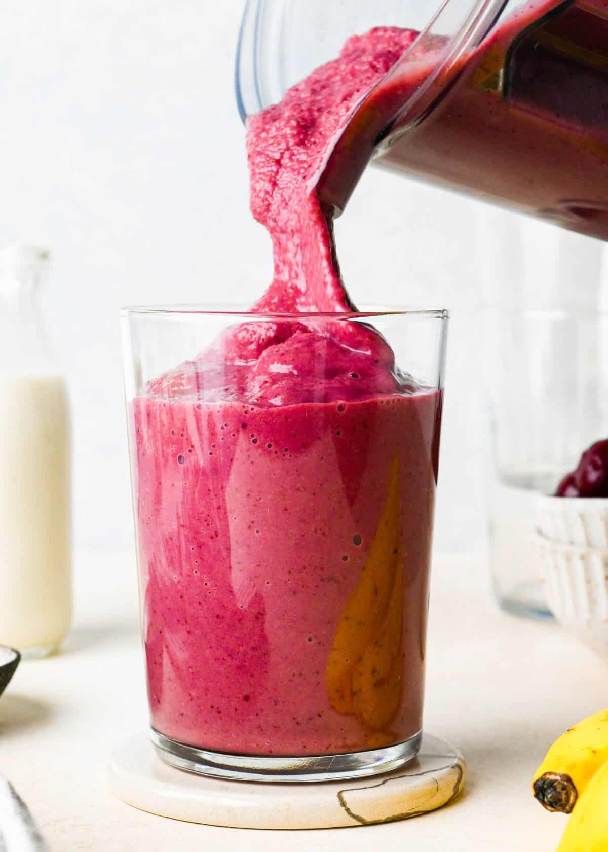 banana cherry smoothie recipe being poured from a blending container into a glass, it's thick texture is visible. there is a glass of milk, bowl of cherries and banana in the background.