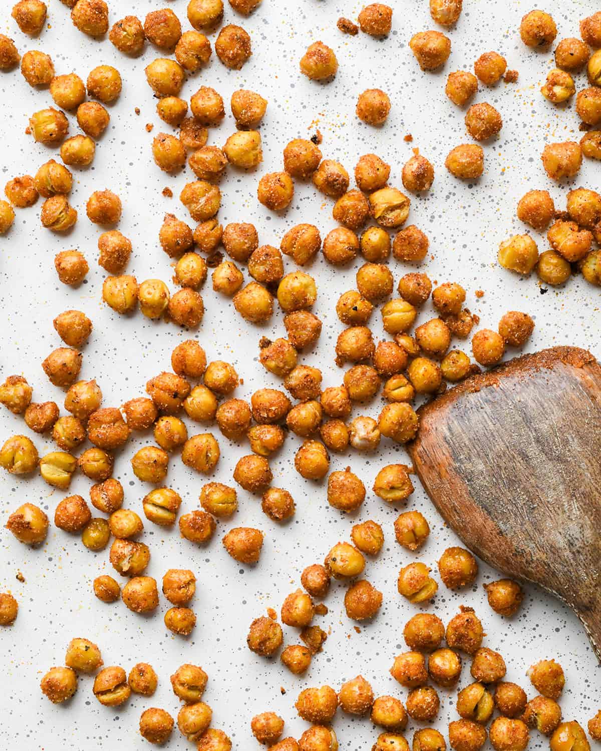 making roasted chickpeas recipe - spice mixture stirred onto the chickpeas on a white baking sheet with a wooden spatula