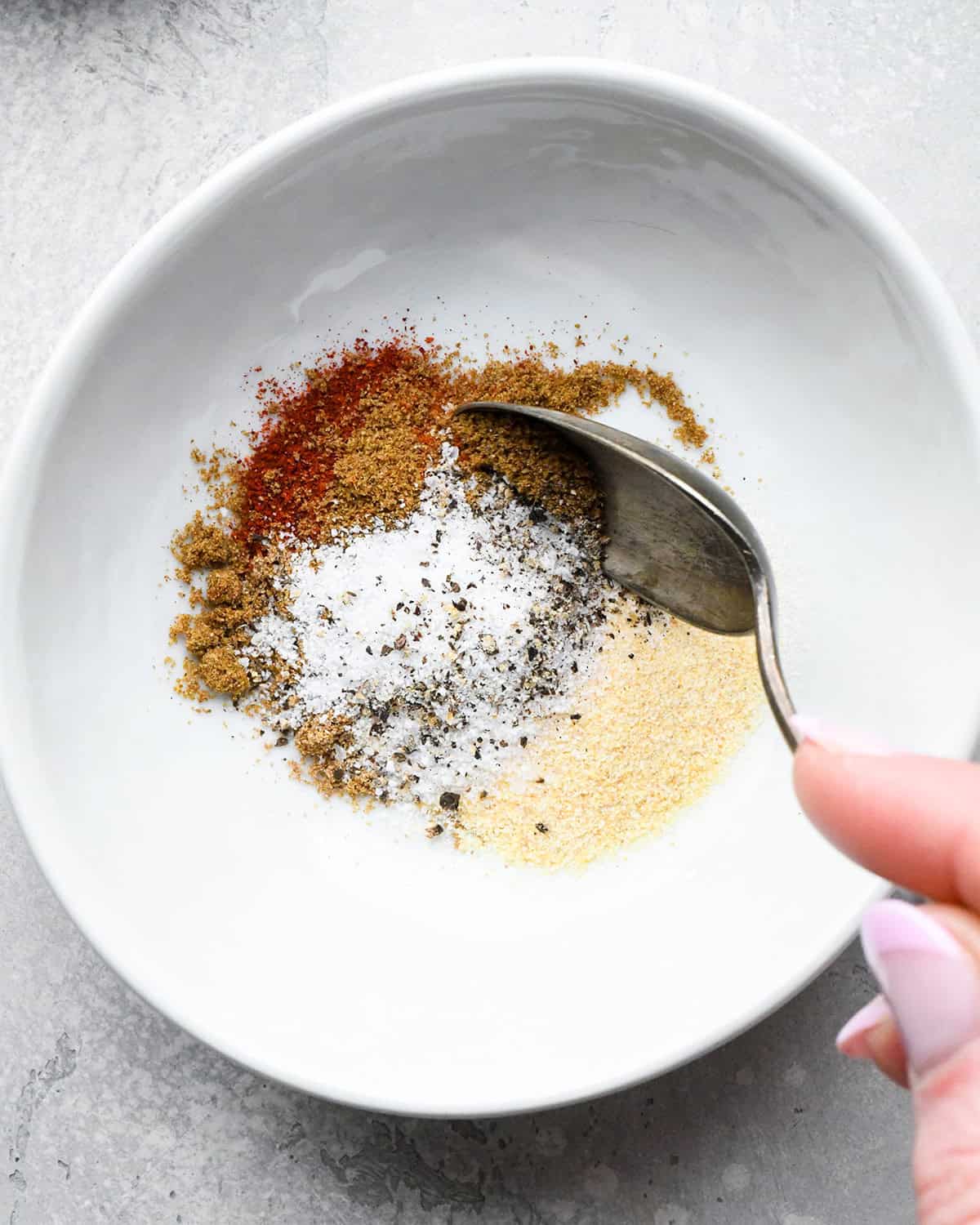 making roasted chickpeas recipe - garlic powder, onion powder, paprika, cumin, coriander, salt and pepper in a small white bowl being stirred with a metal spoon