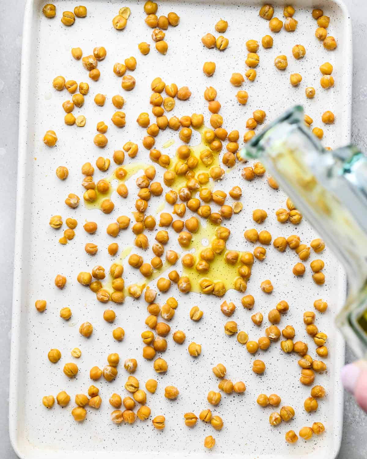 making roasted chickpeas recipe - olive oil being poured over partially-roasted chickpeas on a white baking sheet