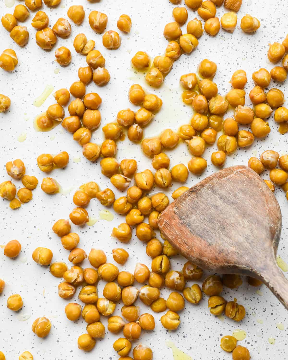 making roasted chickpeas recipe - a wooden spatula stirring the olive oil into the partially roasted chickpeas on a white baking sheet