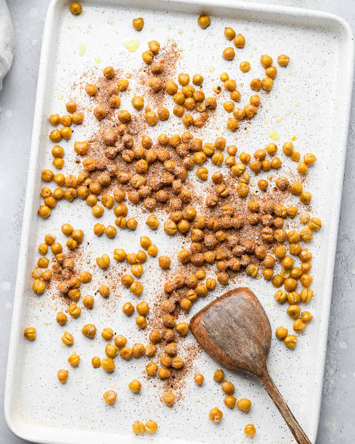 making roasted chickpeas recipe - spice mixture added to the olive-oil coated chickpeas on a white baking sheet with a wooden spatula also on the baking sheet