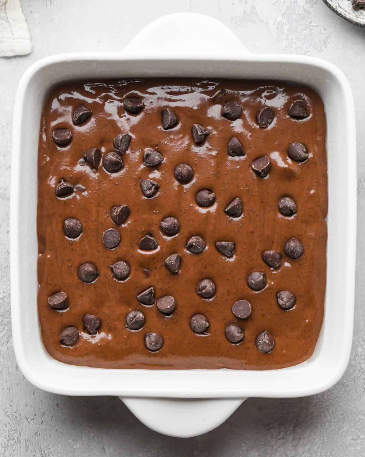 black bean brownies batter spread evenly into a white ceramic square baking dish before baking topped with chocolate chips