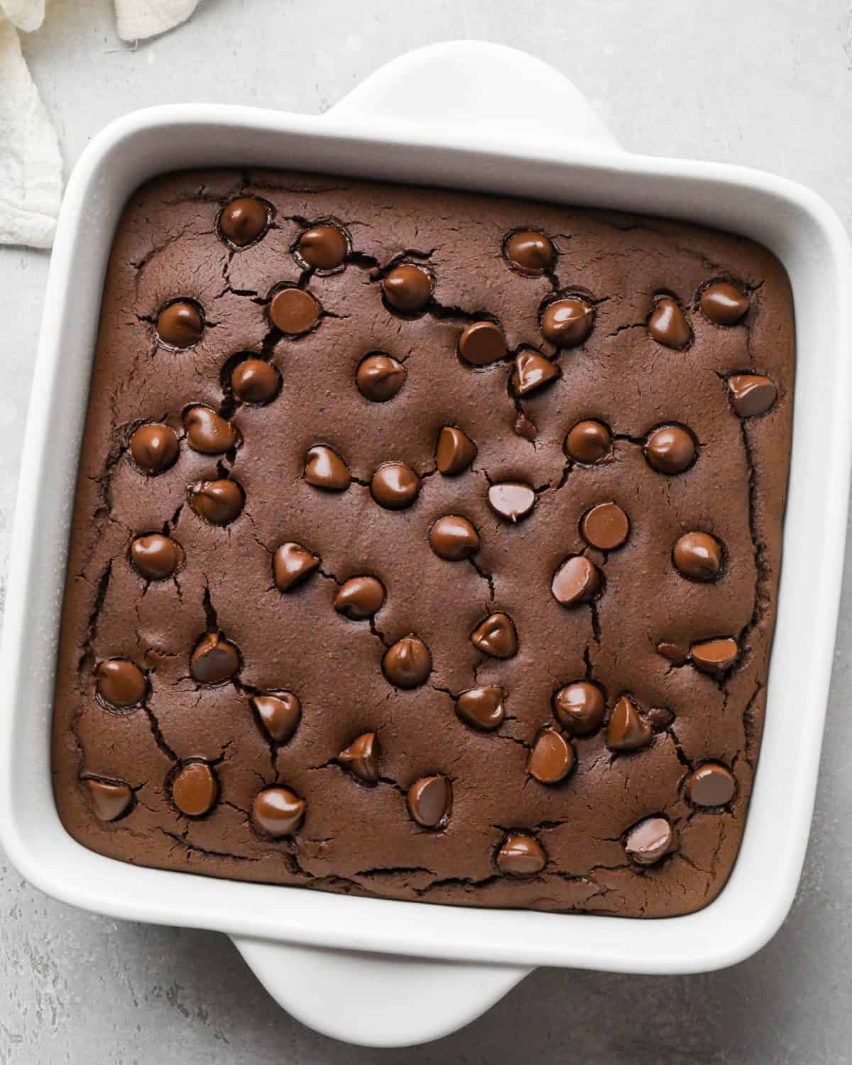 black bean brownies in a white square ceramic baking dish after baking. They have crackly tops and melty chocolate chips on top.