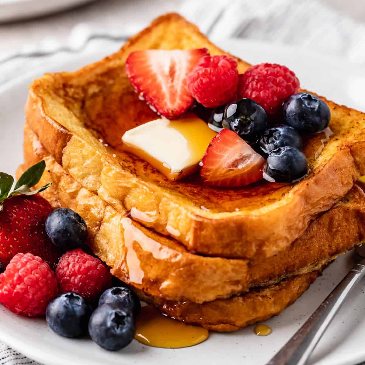a stack of 3 pieces of French Toast with butter, syrup and berries