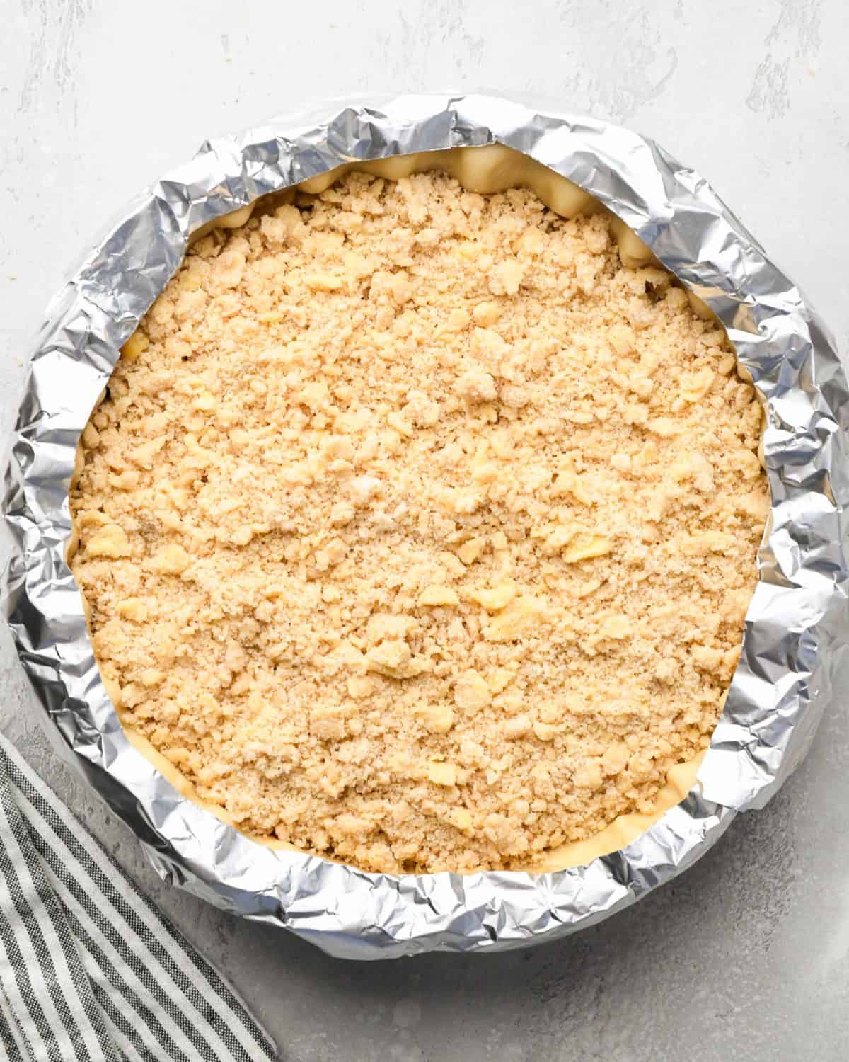 assembled apple crumble pie with a fluted pie crust and a crumb topping with a foil ring around the outer crust
