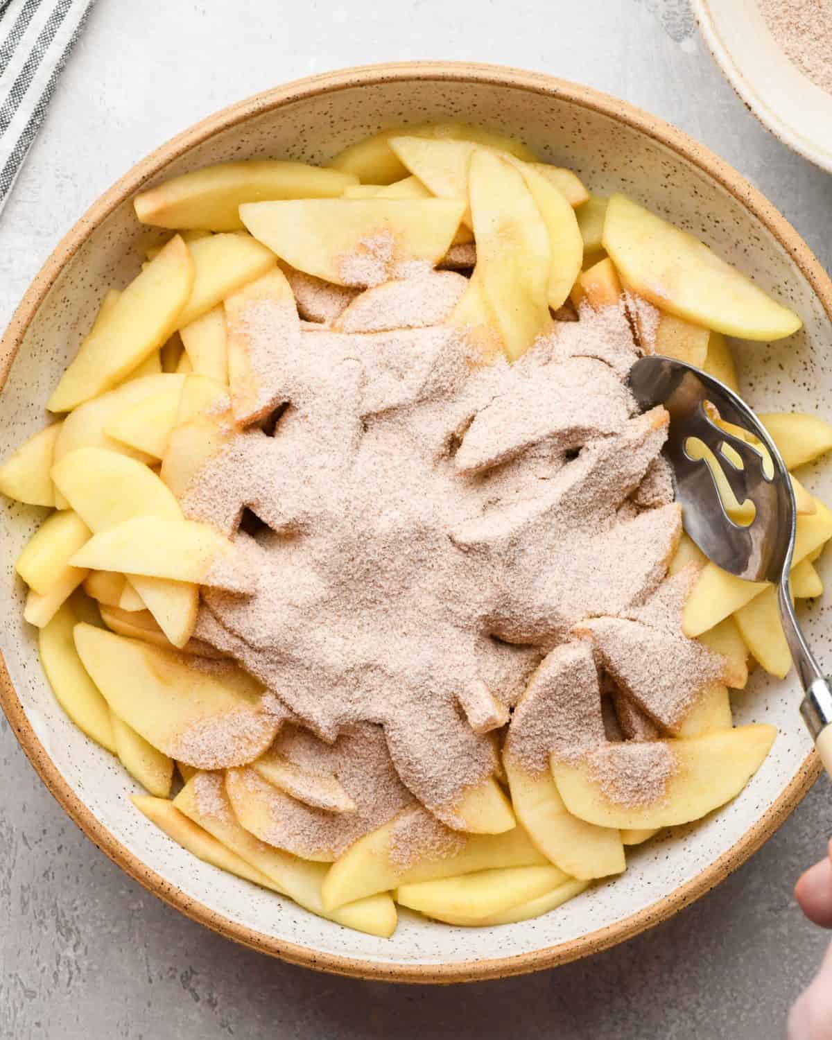 making apple crumb pie - drained apples in a white ceramic mixing bowl with the dry filling ingredient mixture added to the top before mixing. There's a metal spoon in the bowl.