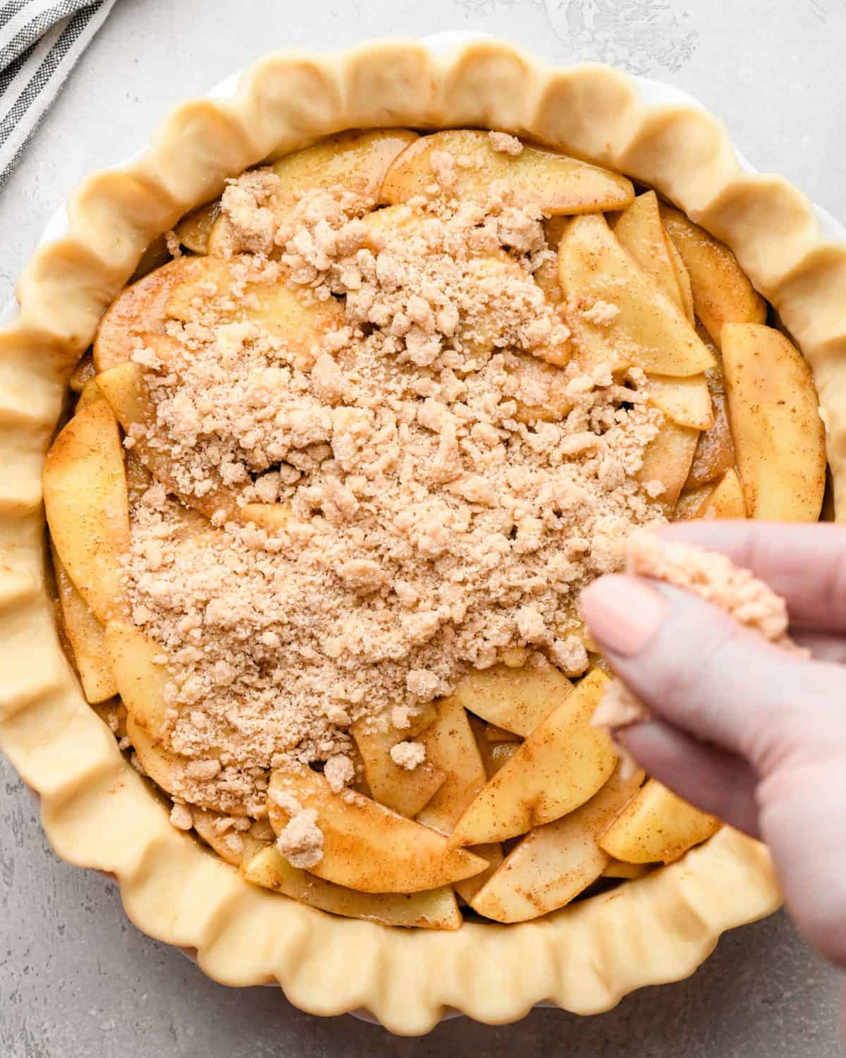 assembling apple crumb pie - a hand sprinkling the crumble topping for apple pie over the apple filling inside a fluted pie crust in a white pie dish on a gray surface. There's a striped towel in the top left corner