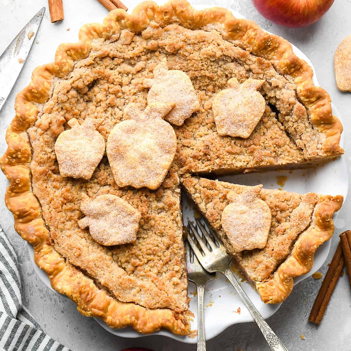 apple crumble pie in a white pie dish with 6 pie crust apples coated in cinnamon sugar on top. There is a slice cut out of the pie and it's in the pie dish with 2 forks. The pie dish is surrounded by apples, pie crust apples and cinnamon sticks.