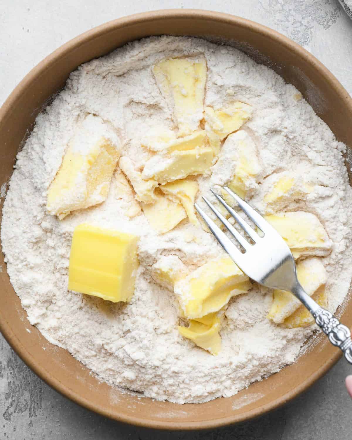 making crumble topping for apple pie - cubes of butter added to the dry ingredients in a brown mixing bowl. The butter is being cut in with a silver fork.