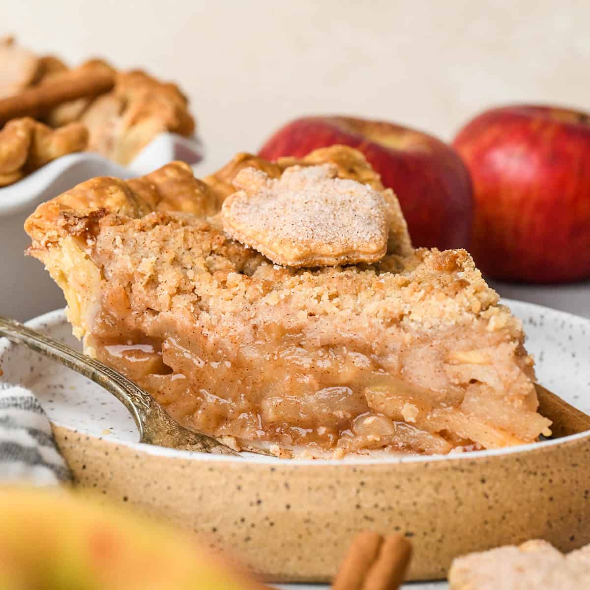 side view of a slice of apple crumb pie on a ceramic plate topped with a pie crust apple. The moist but thick apple filling is visible. There's a fork on the plate, the rest of the pie and apples in the background.