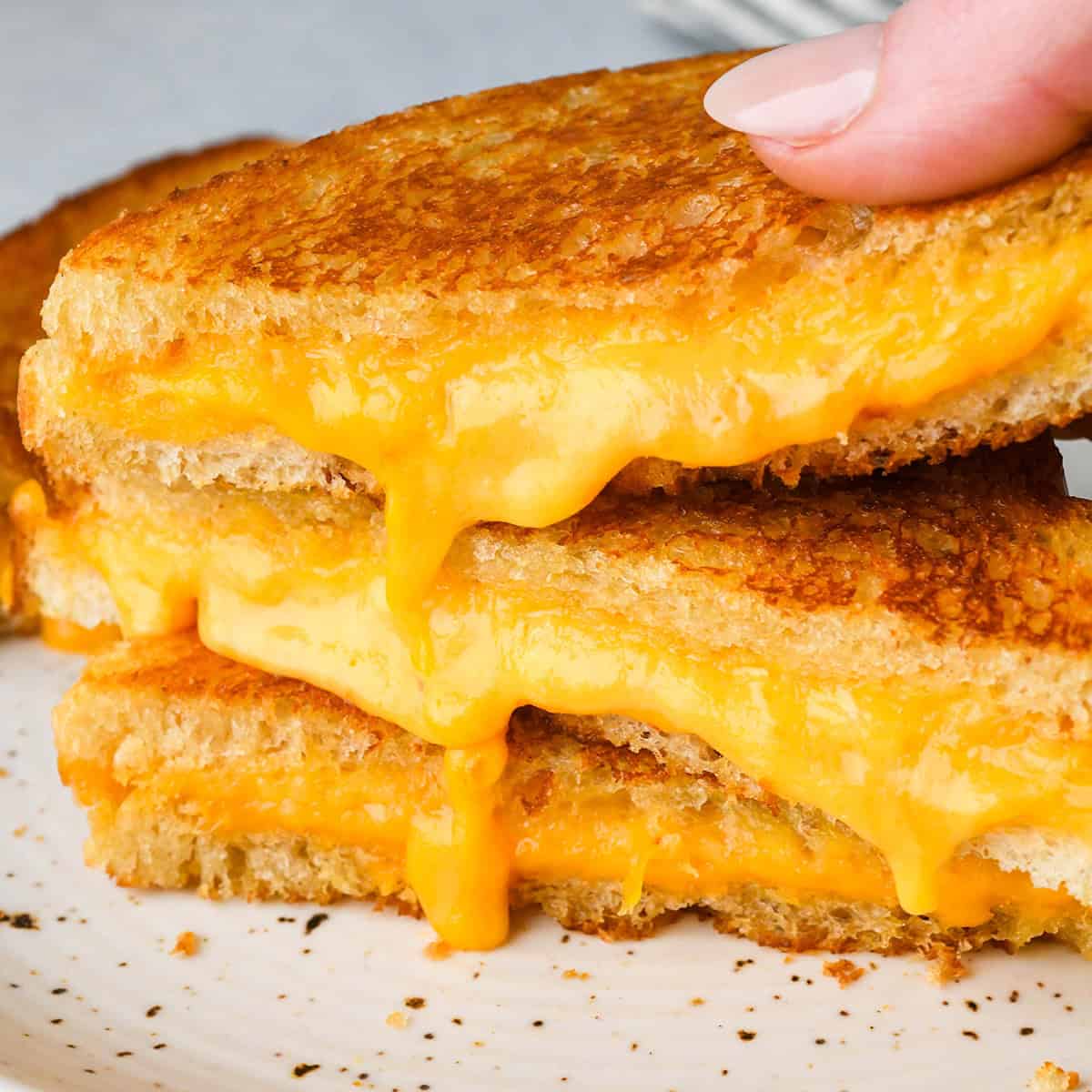 close up photo of a stack of three halves of a grilled cheese sandwich on a white ceramic plate with golden brown bread and melty cheese dripping down the front of the stack a hand is picking up the top half.