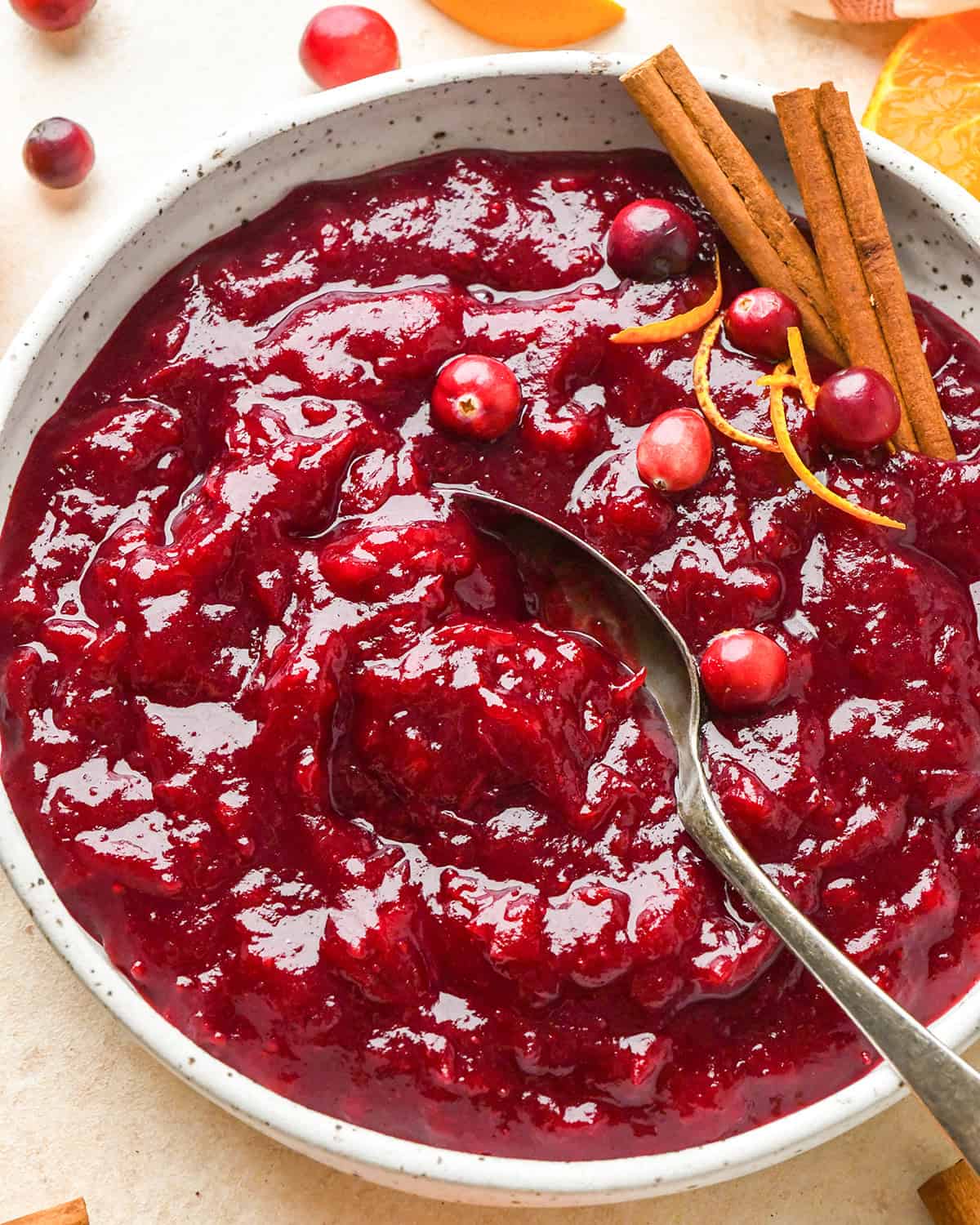 a silver spoon taking a scoop of homemade cranberry sauce with orange juice in a ceramic bowl. You can really see the thick texture. It's topped with whole cranberries, two cinnamon sticks and orange peel. 