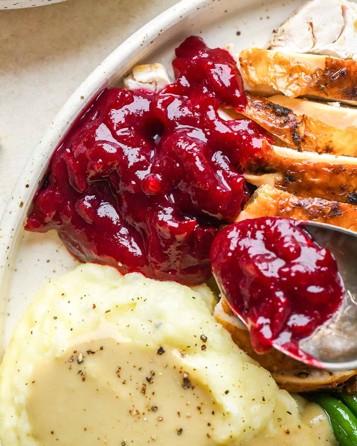 homemade cranberry sauce recipe on a white ceramic plate with turkey, mashed potatoes, green beans and stuffing and a silver spoon with a scoop of cranberry sauce on it hovering over the turkey