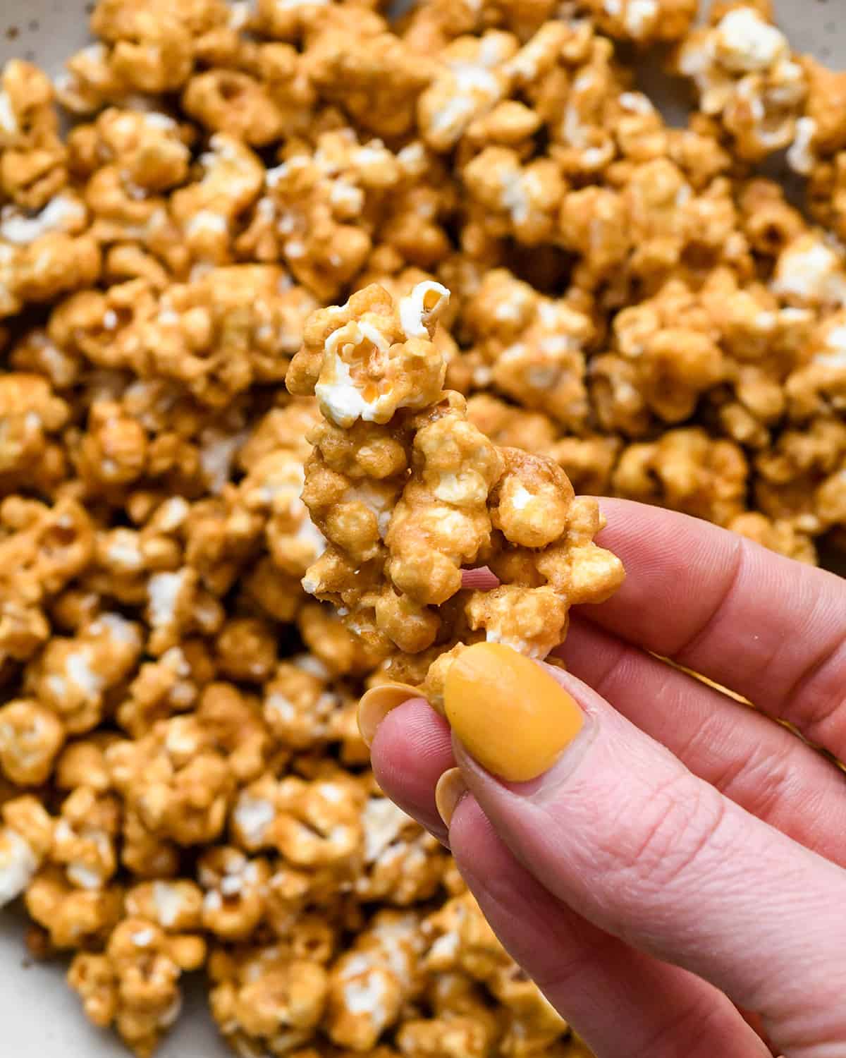 a hand holding a piece of Caramel Popcorn over a bowl of Caramel popcorn