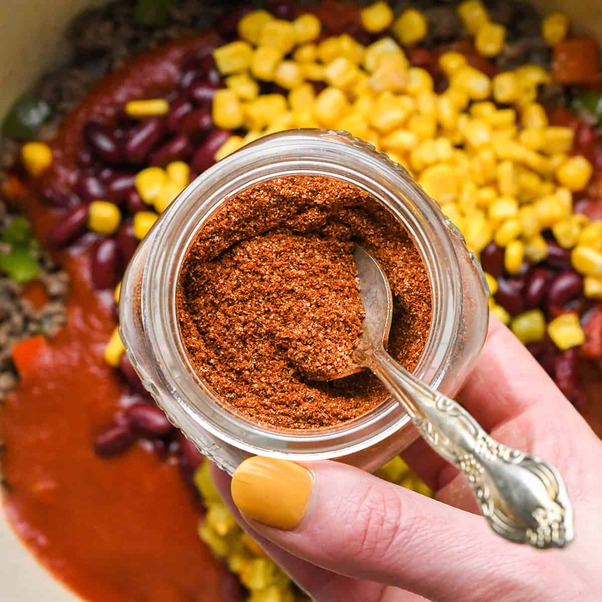 Homemade Chili Seasoning in a glass jar being held over a pot of chili before adding. 