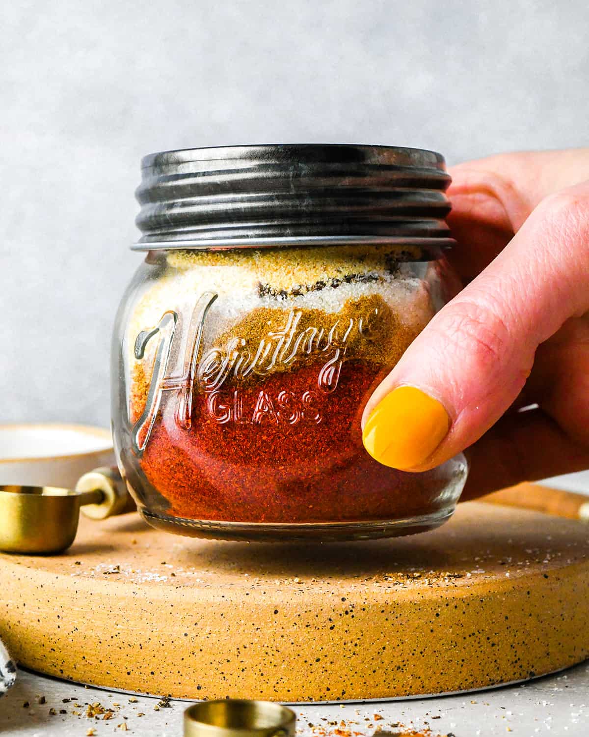 a hand holding a glass jar with chili seasoning ingredients inside and a lid on before shaking