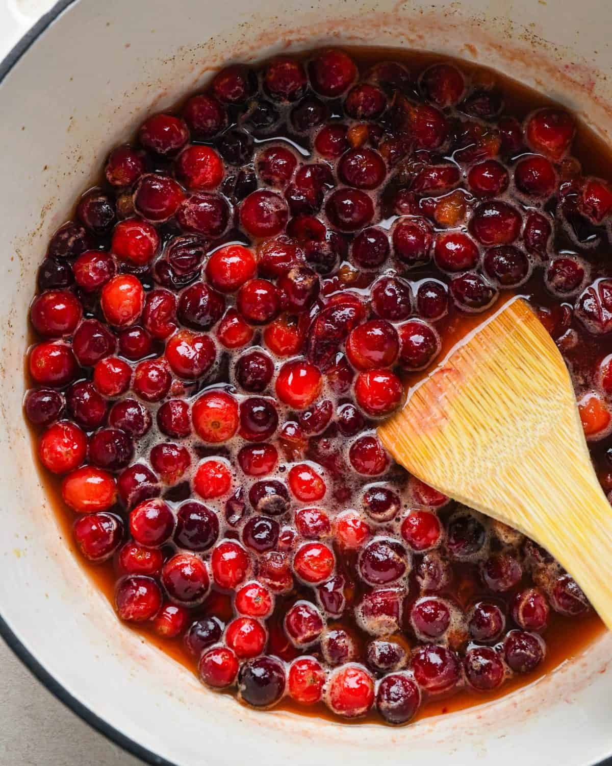 how to make cranberry sauce - a wooden spatula stirring the cranberry sauce mixture as it cooks in a white pot with a black rim