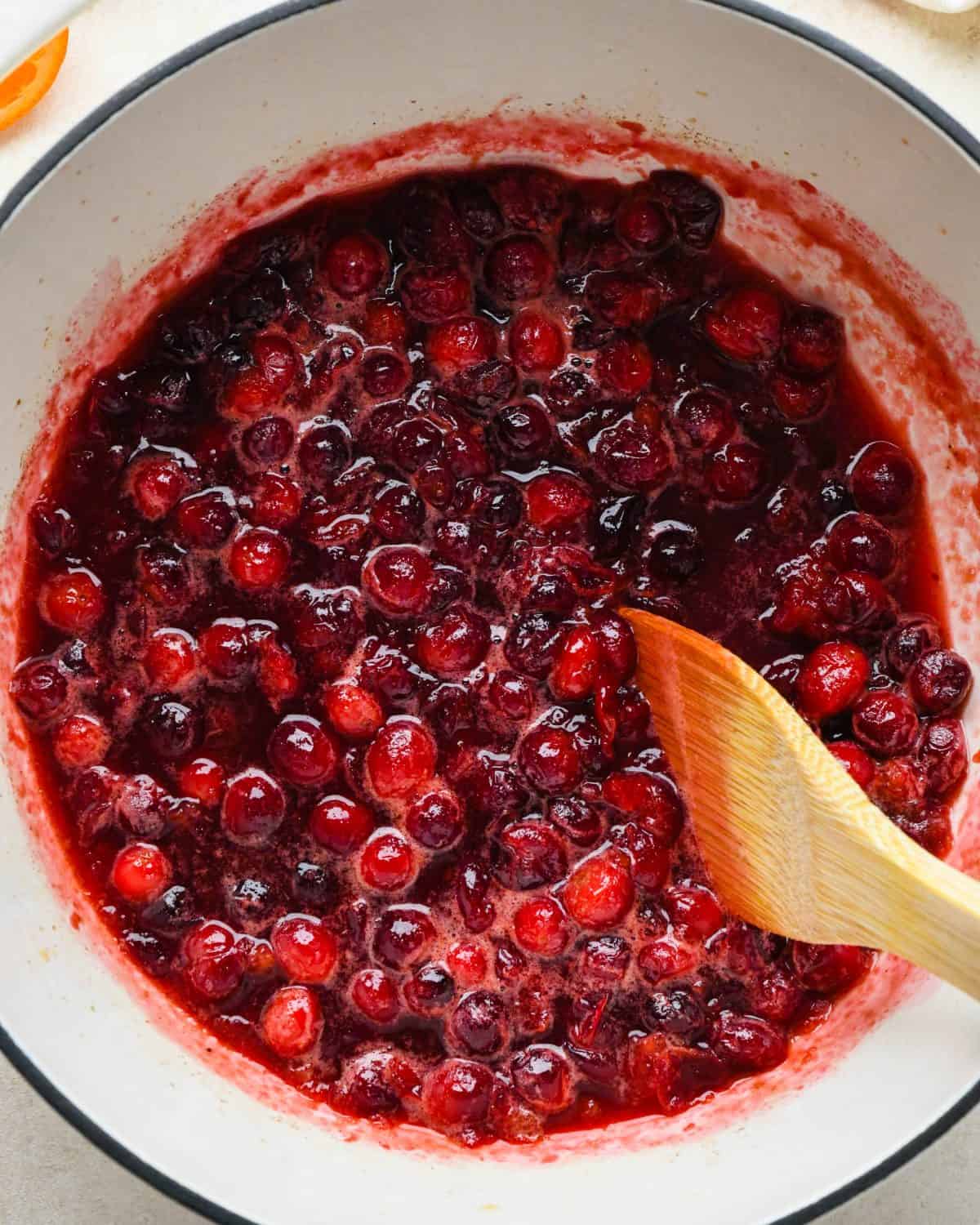 how to make cranberry sauce - a wooden spatula stirring the cranberry sauce mixture as it cooks in a white pot with a black rim.  more of the berries have popped.