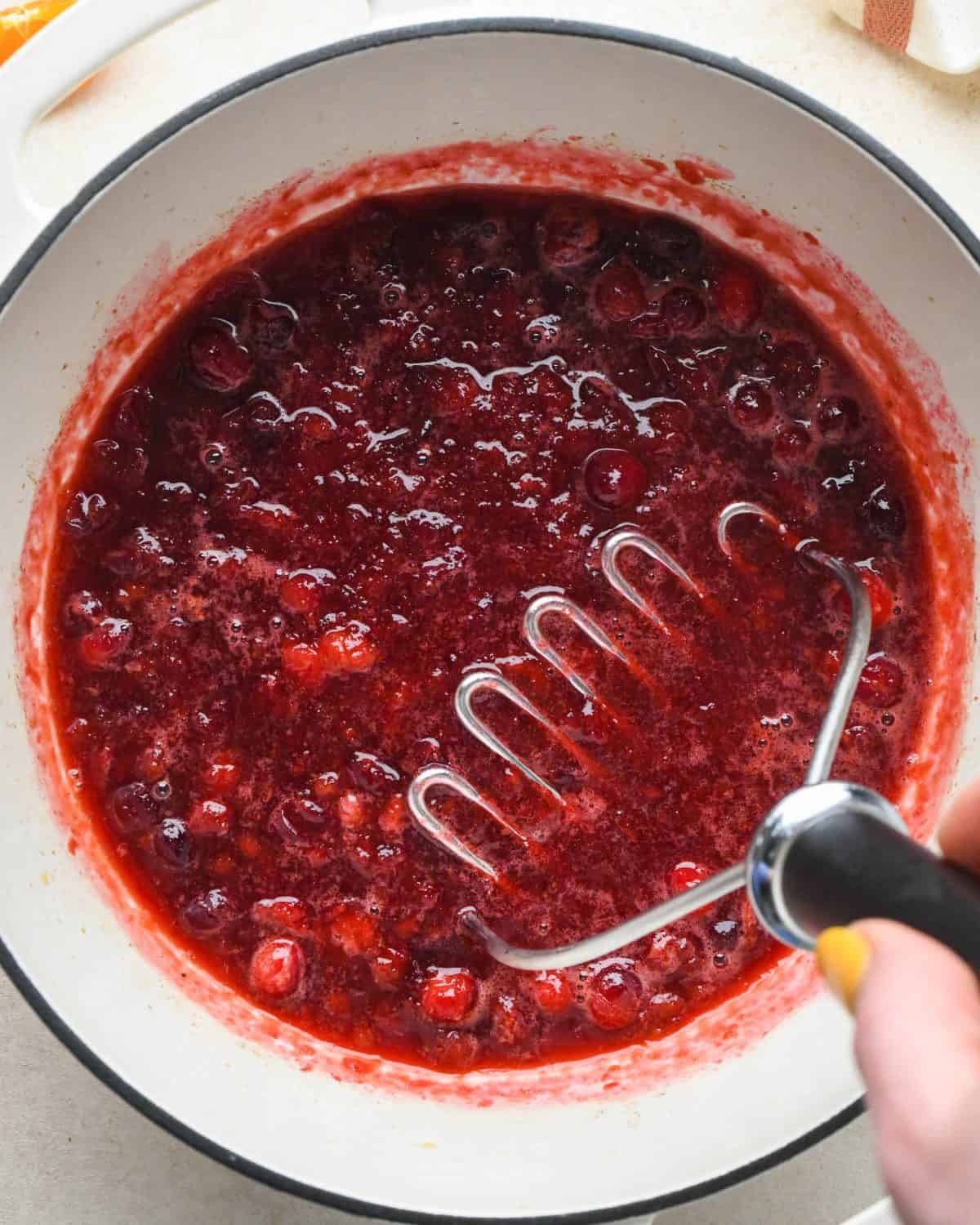 how to make cranberry sauce - a potato masher being used to mash the cranberry sauce mixture in a white pot with a black rim