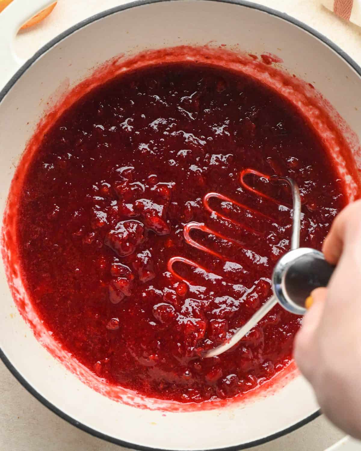 how to make cranberry sauce - a potato masher being used to mash the cranberry sauce mixture in a white pot with a black rim