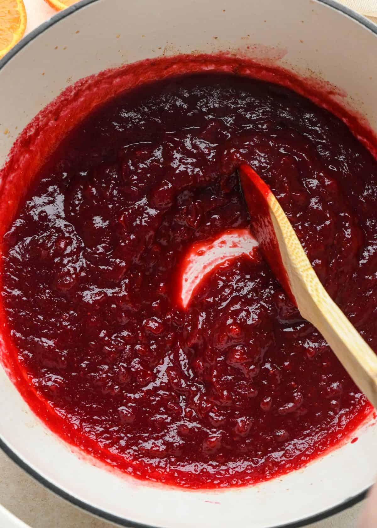  a wooden spatula stirring the cranberry sauce with orange juice as it cools, showing the thick texture of the sauce. 