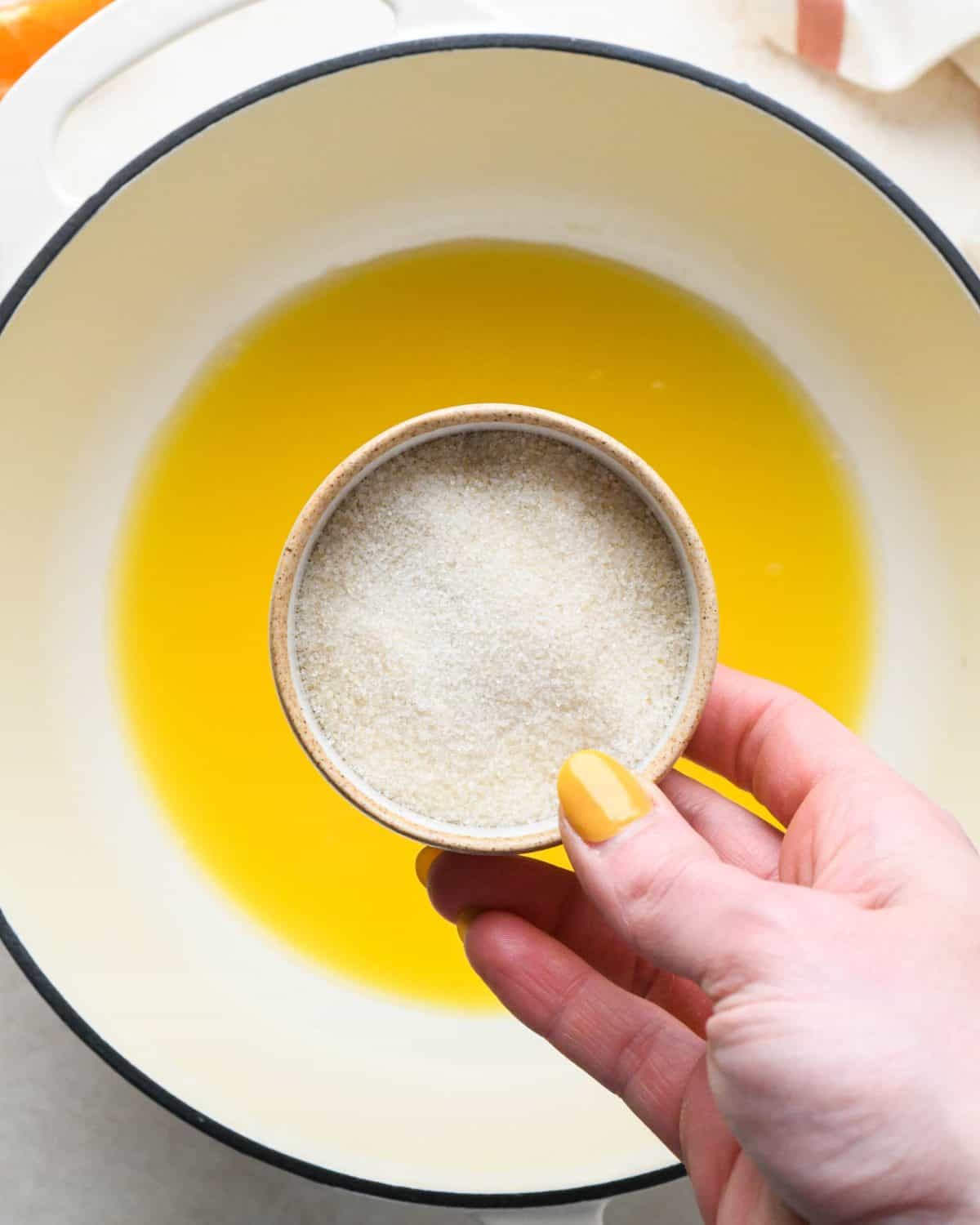 how to make cranberry sauce - a bowl of granulated sugar being held over a white pot with a black rim with orange juice in it