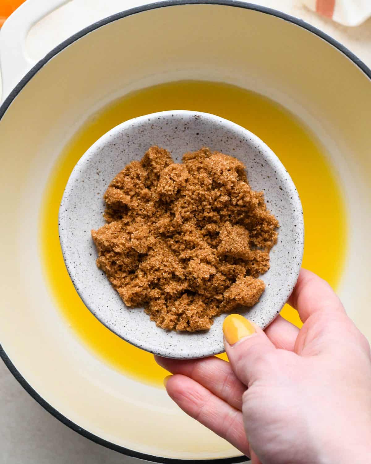 how to make cranberry sauce - a bowl of brown sugar being held over a white pot with a black rim with orange juice in it