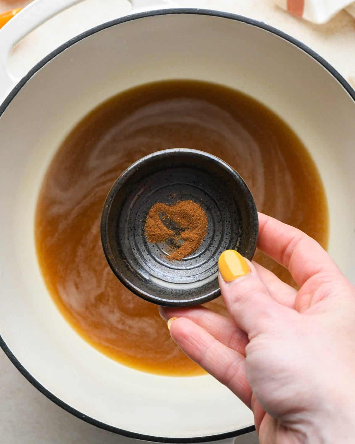 how to make cranberry sauce -  a hand holding a small bowl of cinnamon over a white pot with a black rim with the rest of the ingredients in it 