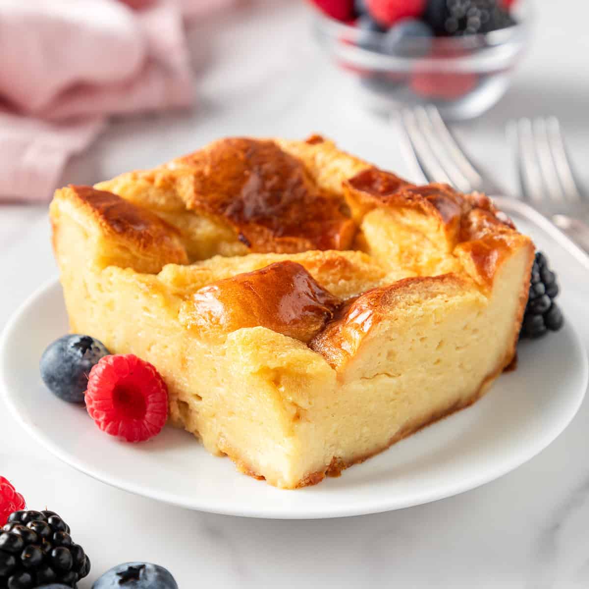 a slice of old fashioned bread pudding on a white plate with a golden brown top and moist interior, surrounded by berries. There are two forks, a bowl of berries and a pink towel in the background.