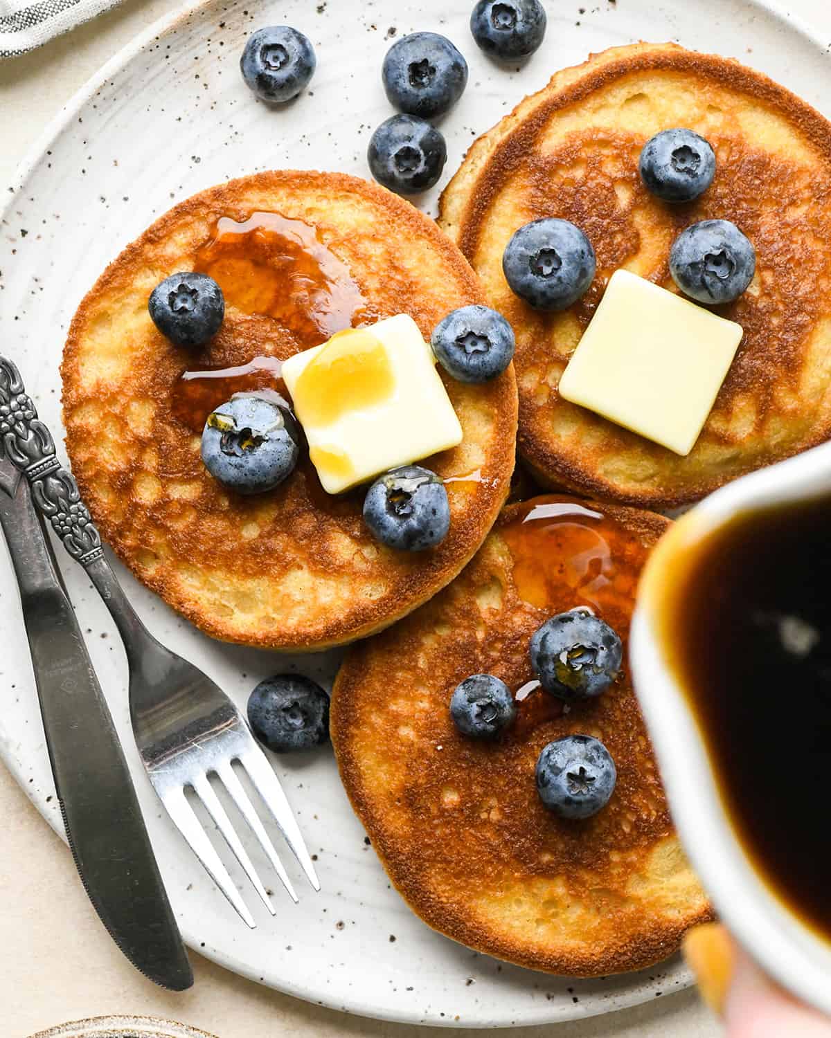 du sirop étant versé sur 3 crêpes à la farine d'amande sur une assiette avec du beurre et des myrtilles