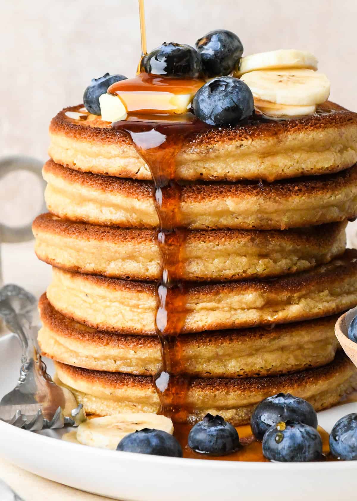 du sirop étant versé sur une pile de 6 crêpes à la farine d'amande avec du beurre et des fruits