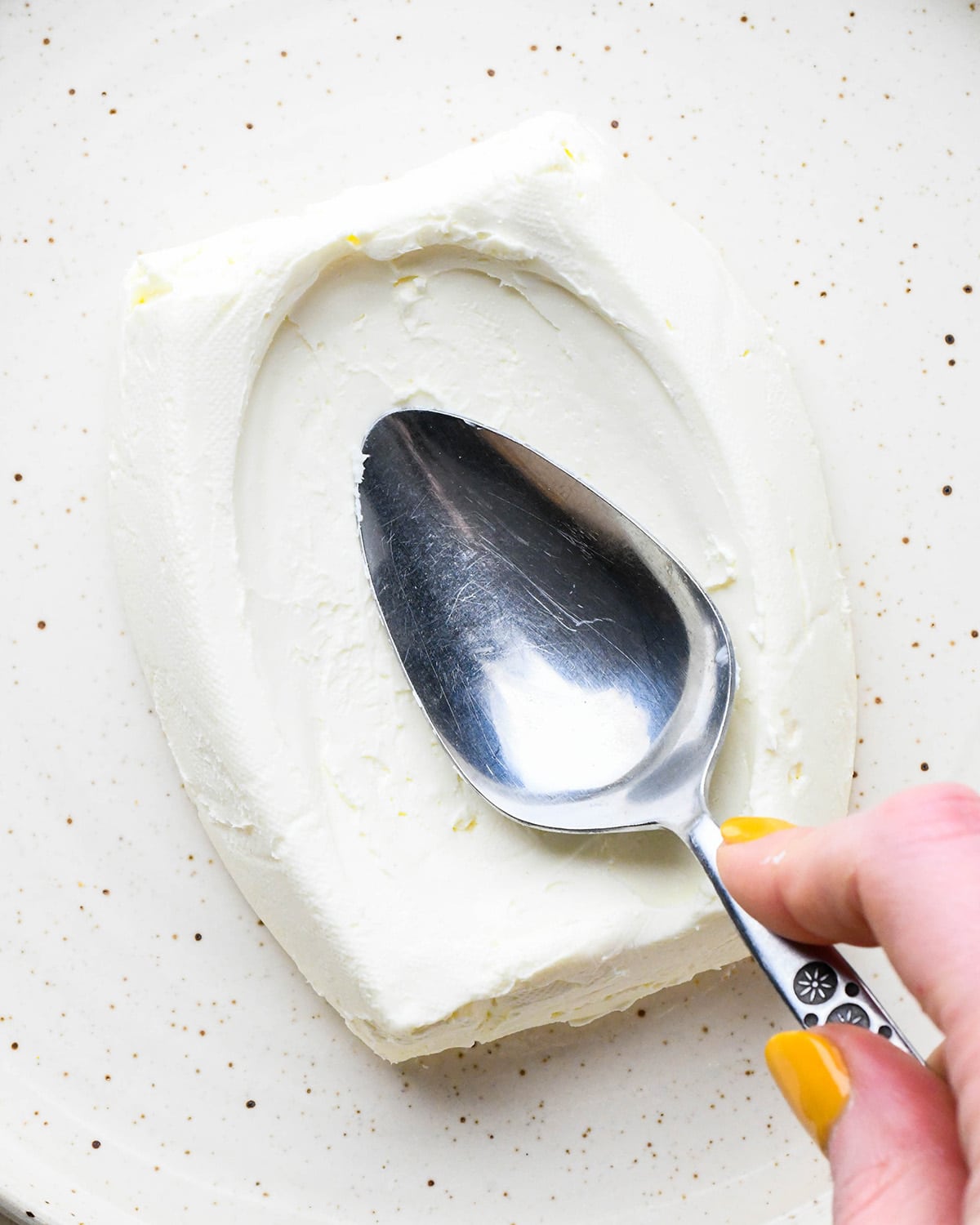 a spoon making a "well" in a block of cream cheese on a plate