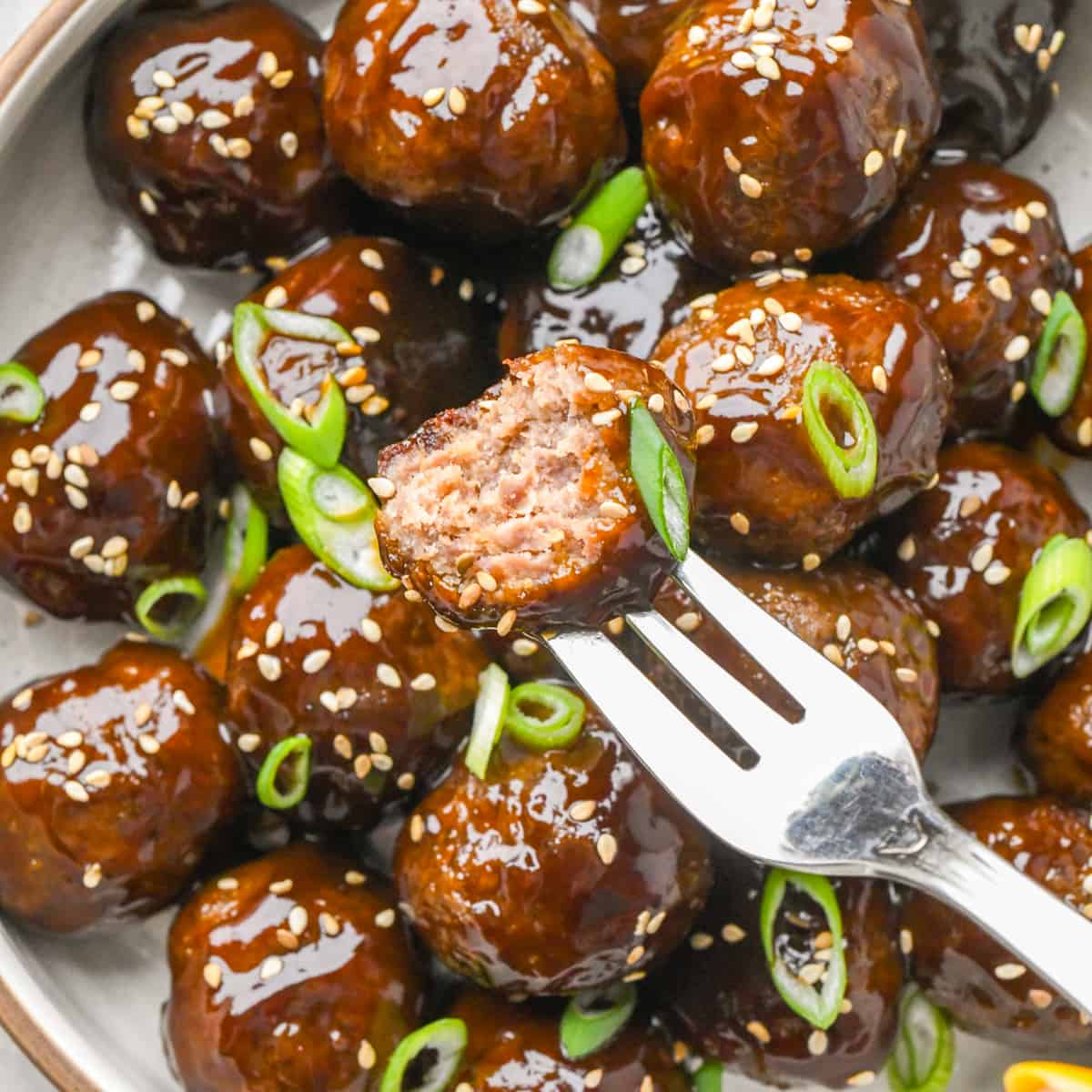 a fork holding an Asian Meatball with a bite taken out of it over a plate of Asian Meatballs garnished with sesame seeds and green onions