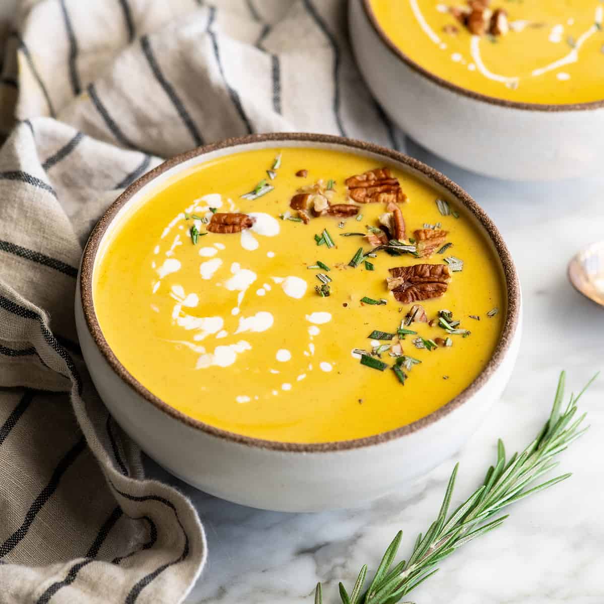 two bowls of Butternut Squash Soup garnished with pecans, cream and rosemary