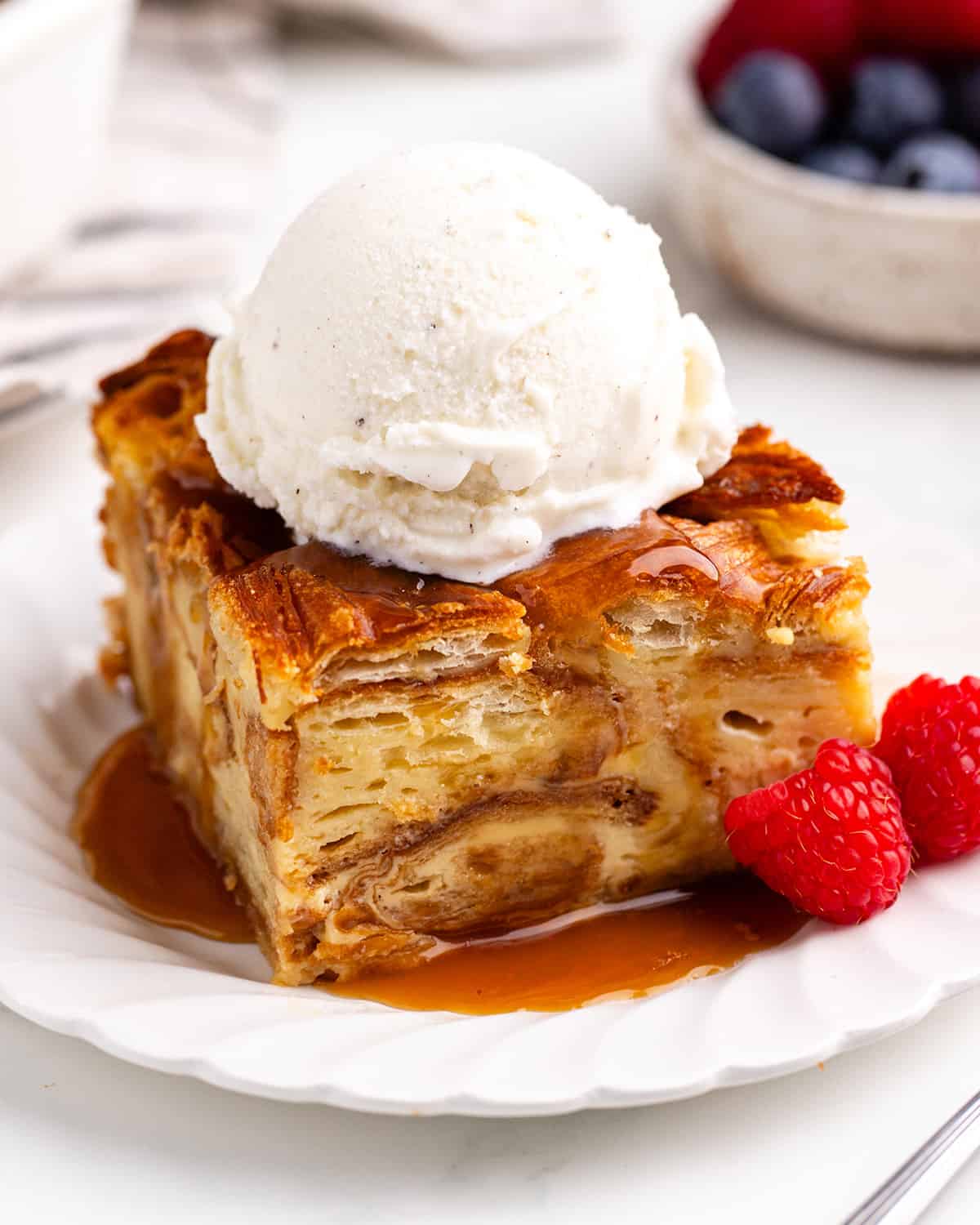 a piece of Croissant Bread Pudding on a plate with vanilla ice cream, caramel sauce and raspberries