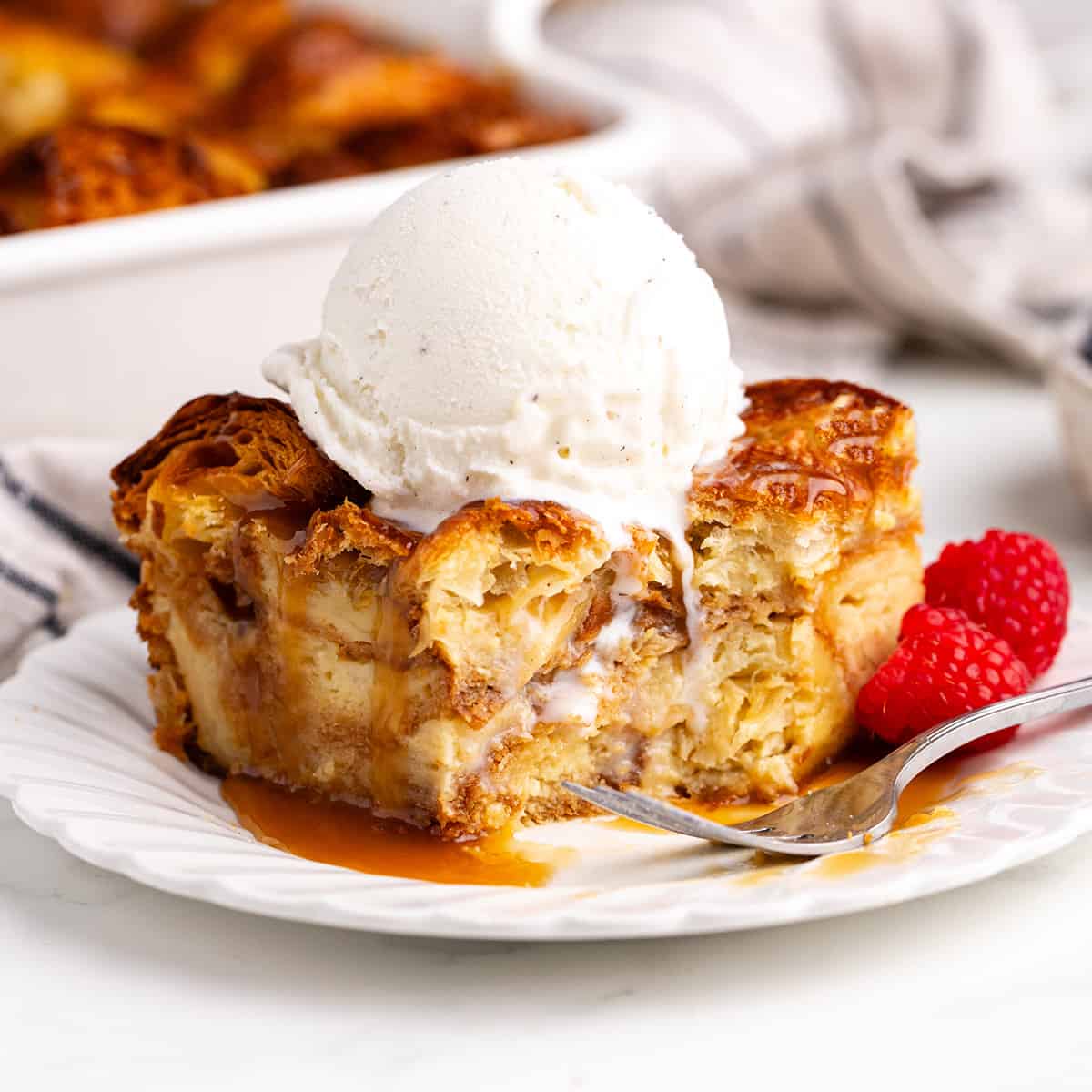 a slice of Croissant Bread Pudding on a plate with vanilla ice cream, caramel sauce and raspberries