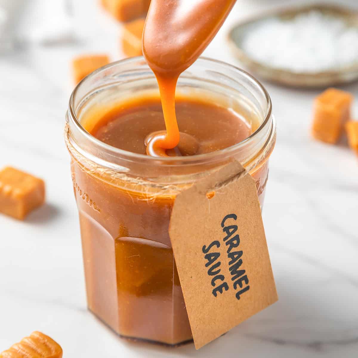 a spoon drizzling some caramel sauce into a jar of caramel sauce with a label