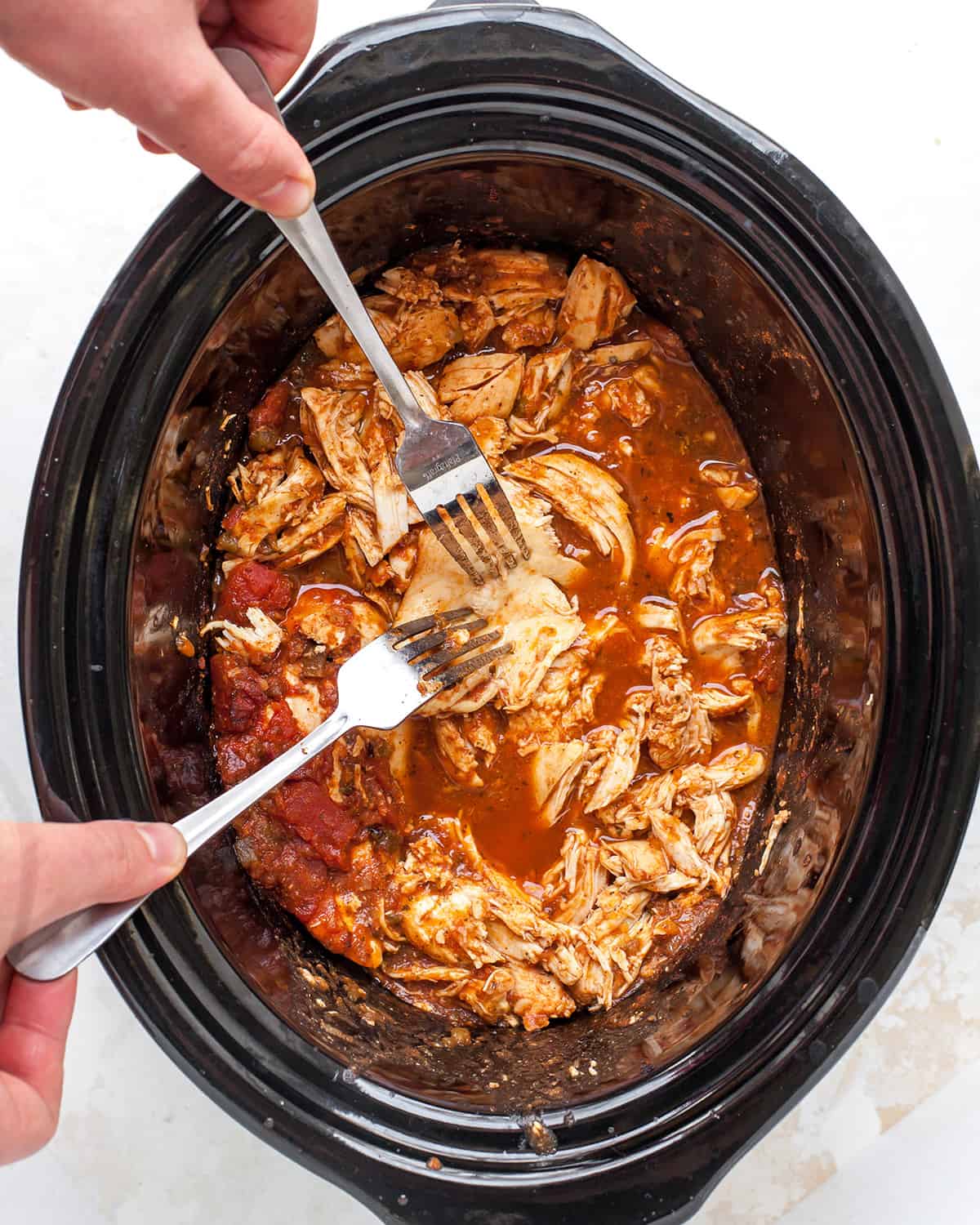 a hand with two forks shredding chicken in a crock pot for shredded chicken tacos