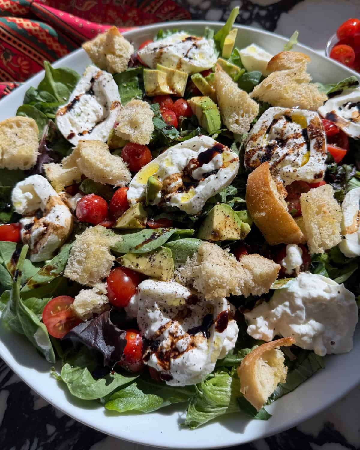 burrata salad on a white serving plate where the burrata cheese has been sliced and placed on top of the salad that contains  halved cherry tomatoes, avocado, fresh basil, and homemade croutons all on spring mix sprinkled with salt and pepper and drizzled with homemade burrata salad dressing. 