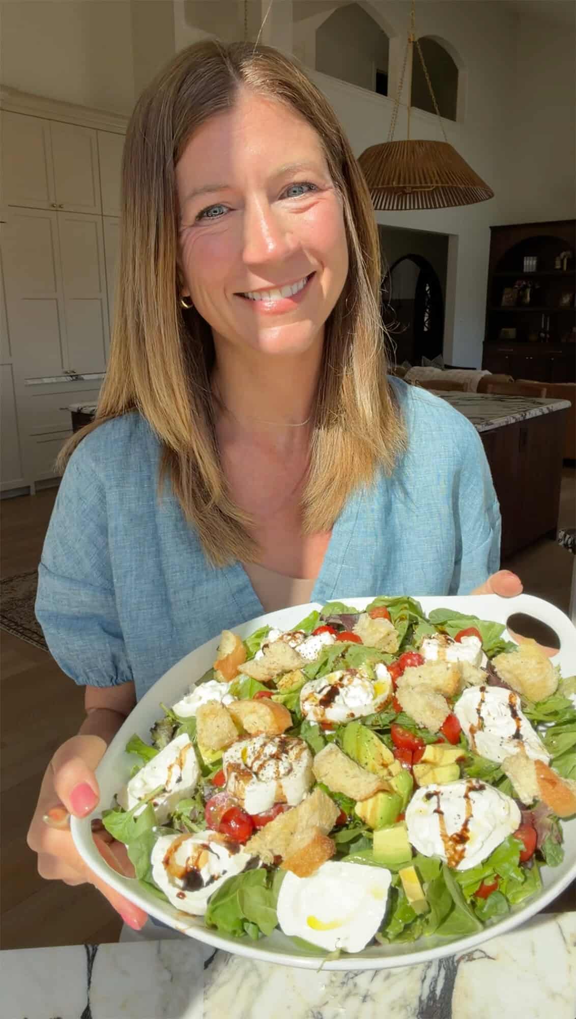 Laura from Joyfoodsunshine holding a white serving plate with burrata salad on it where the burrata cheese has been sliced and placed on top of the salad that contains  halved cherry tomatoes, avocado, fresh basil, and homemade croutons all on spring mix sprinkled with salt and pepper and drizzled with homemade burrata salad dressing. 