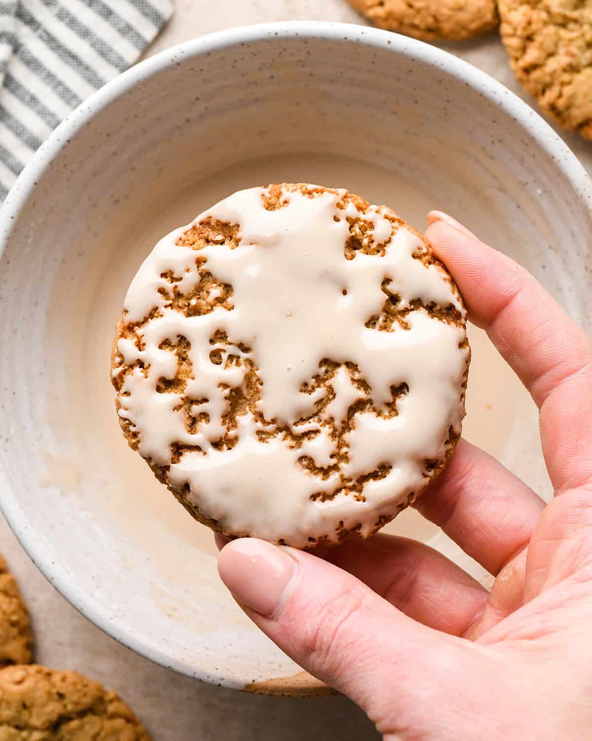 Iced Oatmeal Cookie being held over a bowl of icing after being dipped in it