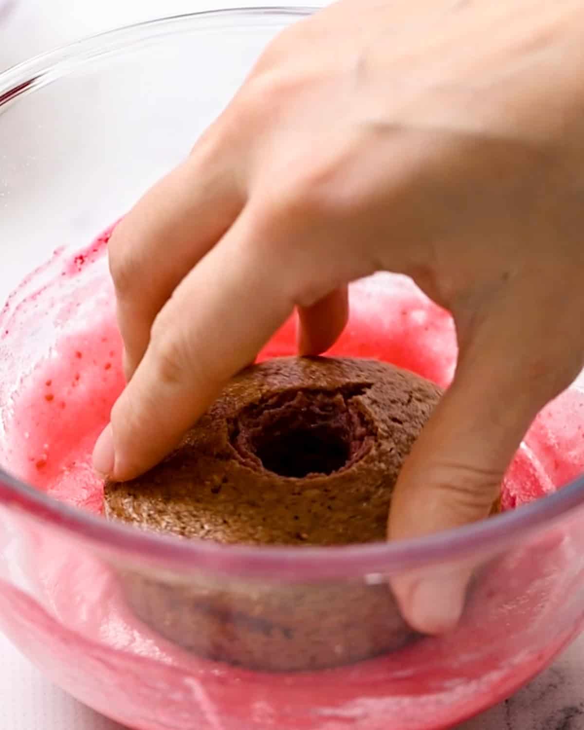 a Baked Raspberry Donut being dipped into raspberry glaze