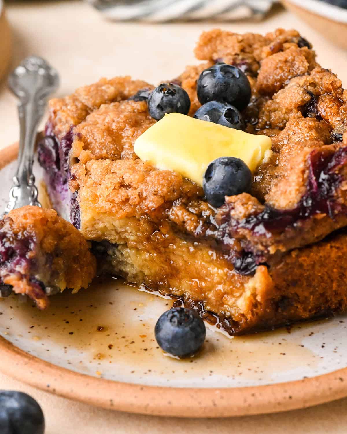 a piece of Blueberry French Toast Casserole  on a plate with butter, syrup and a fork with a bite on it
