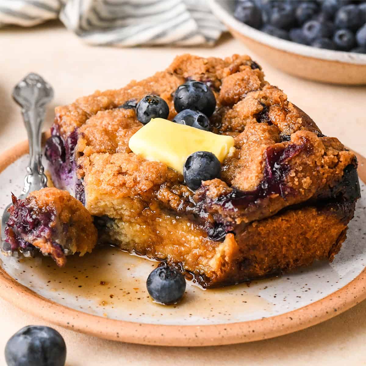 a piece of Blueberry French Toast Casserole  on a plate with butter, syrup and a fork with a bite on it