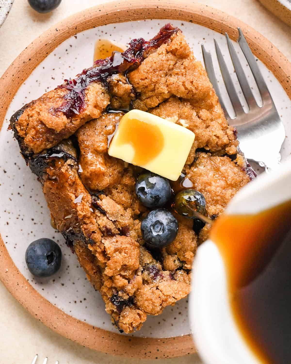 syrup being poured on a piece of Blueberry French Toast Casserole  with butter and blueberries