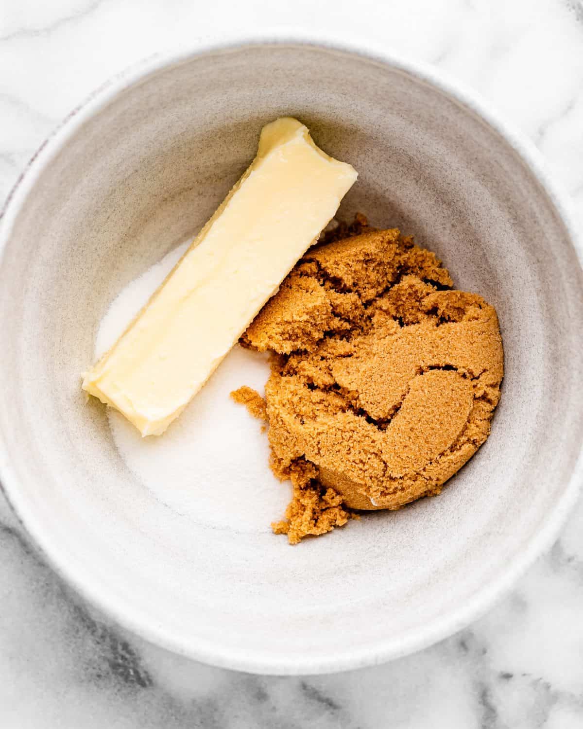 making cherry cookies - a stick of butter, white sugar and brown sugar in a ceramic mixing bowl on a marble surface before mixing
