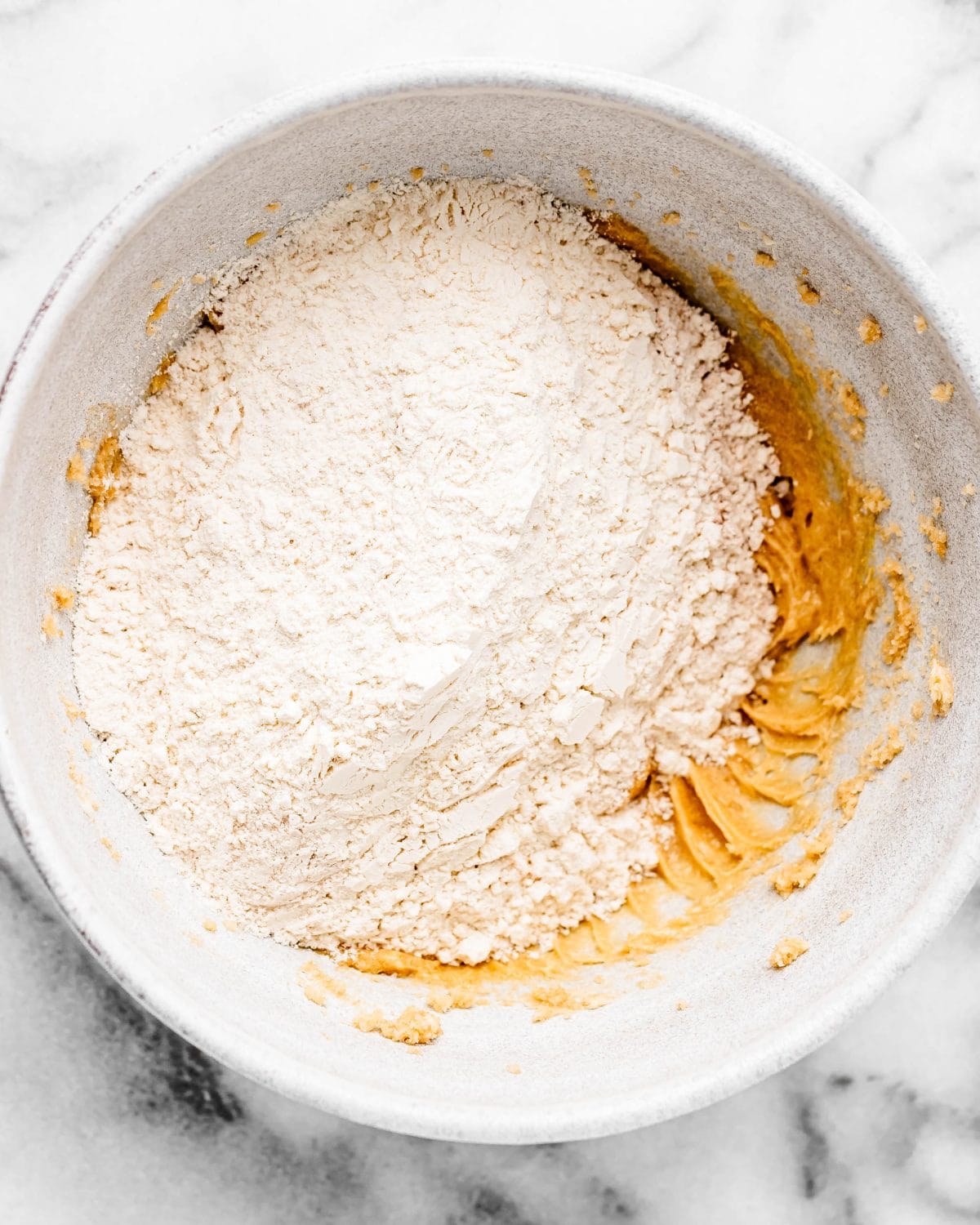 making cherry cookies - dry flour mixture added to the wet ingredient mixture in a white ceramic mixing bowl on a marble surface before mixing