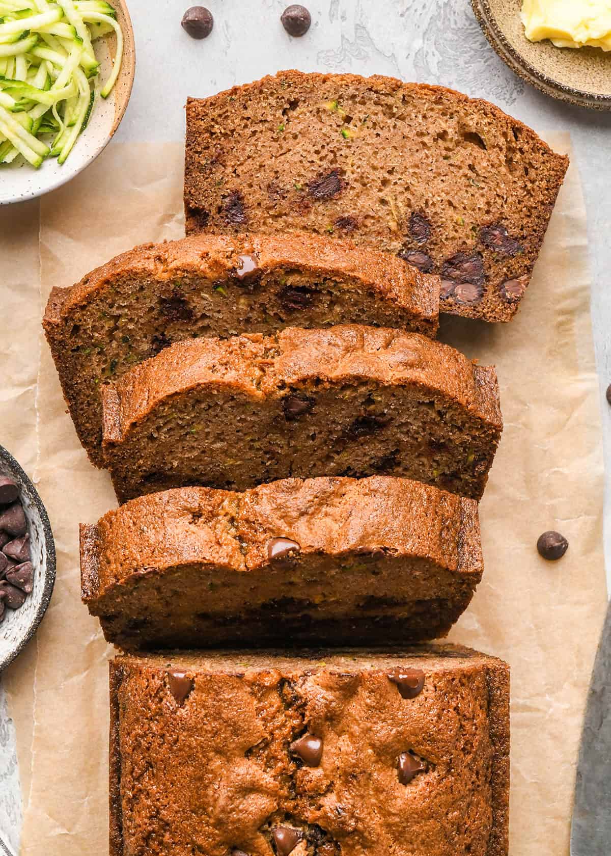 a loaf of Chocolate Chip Zucchini Bread with 4 slices cut out of it on parchment paper with shredded zucchini, chocolate chips and butter in bowls nearby