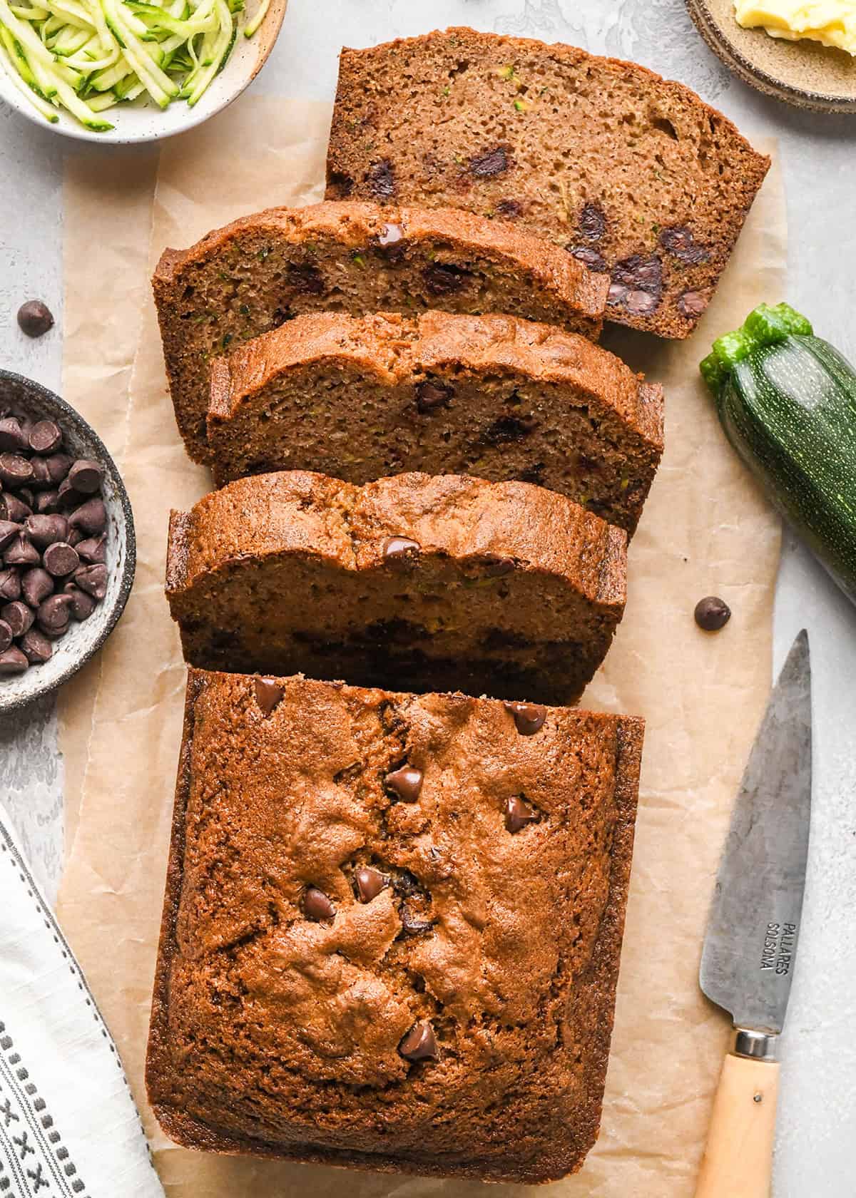 a loaf of Chocolate Chip Zucchini Bread with 4 slices cut out of it on parchment paper with shredded zucchini, chocolate chips and butter in bowls nearby